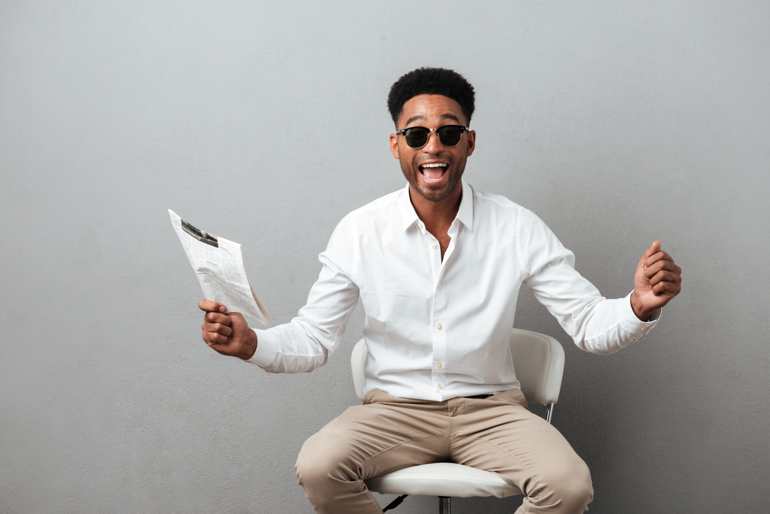 Happy excited african man holding newspaper and celebrating a win while sitting in a chair isolated over gray background
